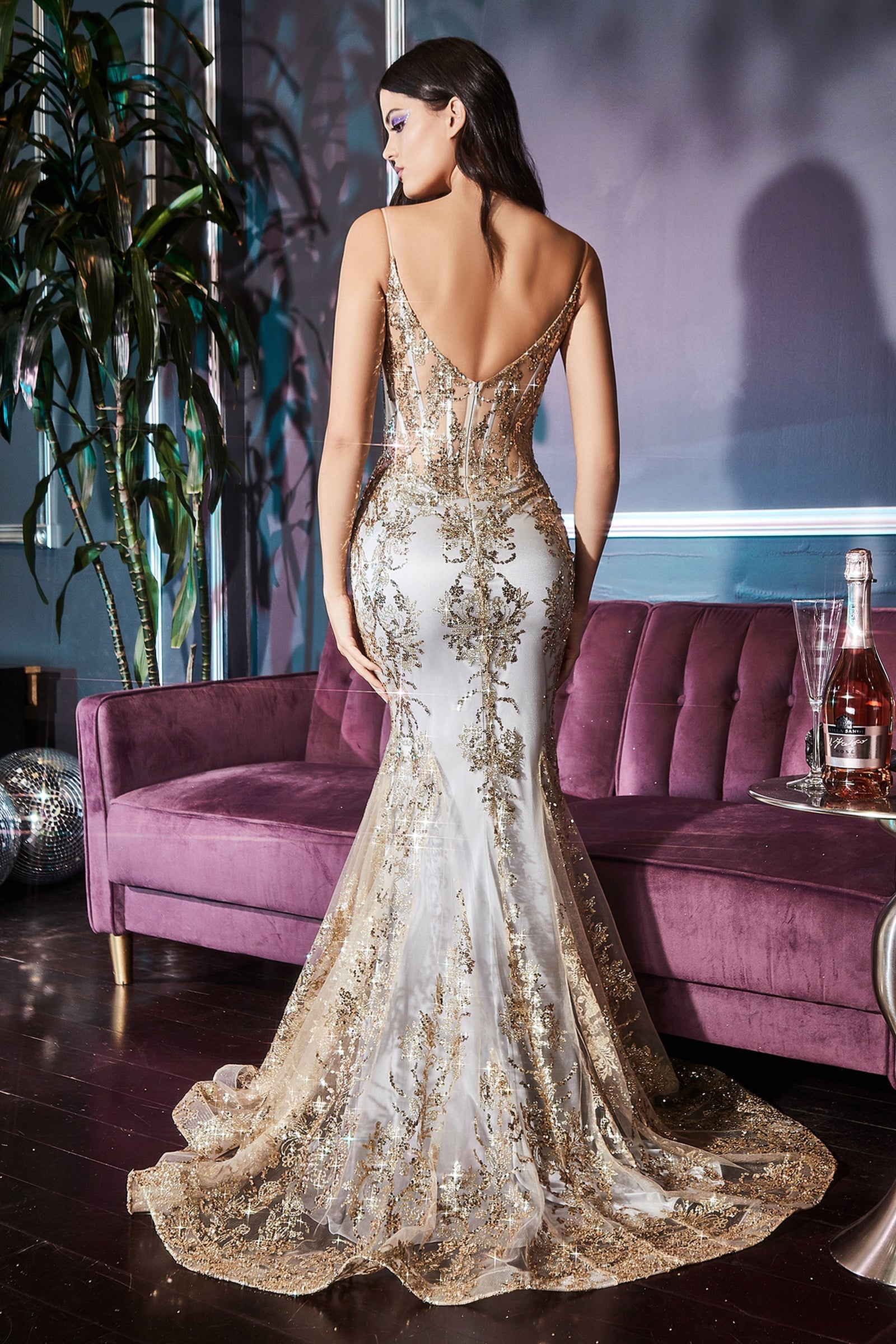 Woman modeling the back of a long gold dress she's wearing standing in front of a velvet purple couch.